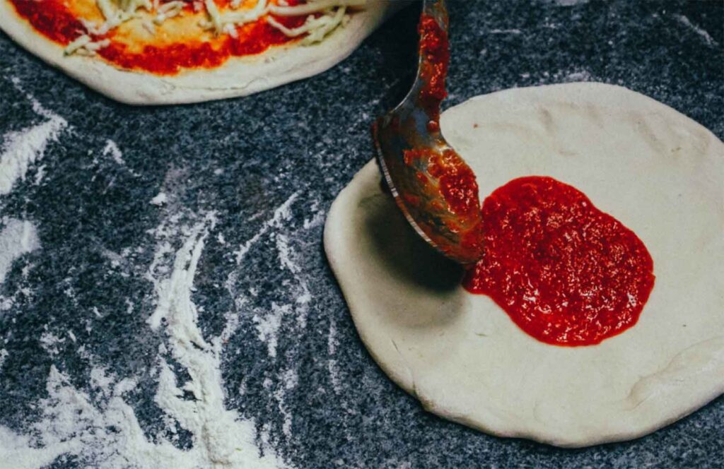 ladling homemade tomato paste on homemade pizza